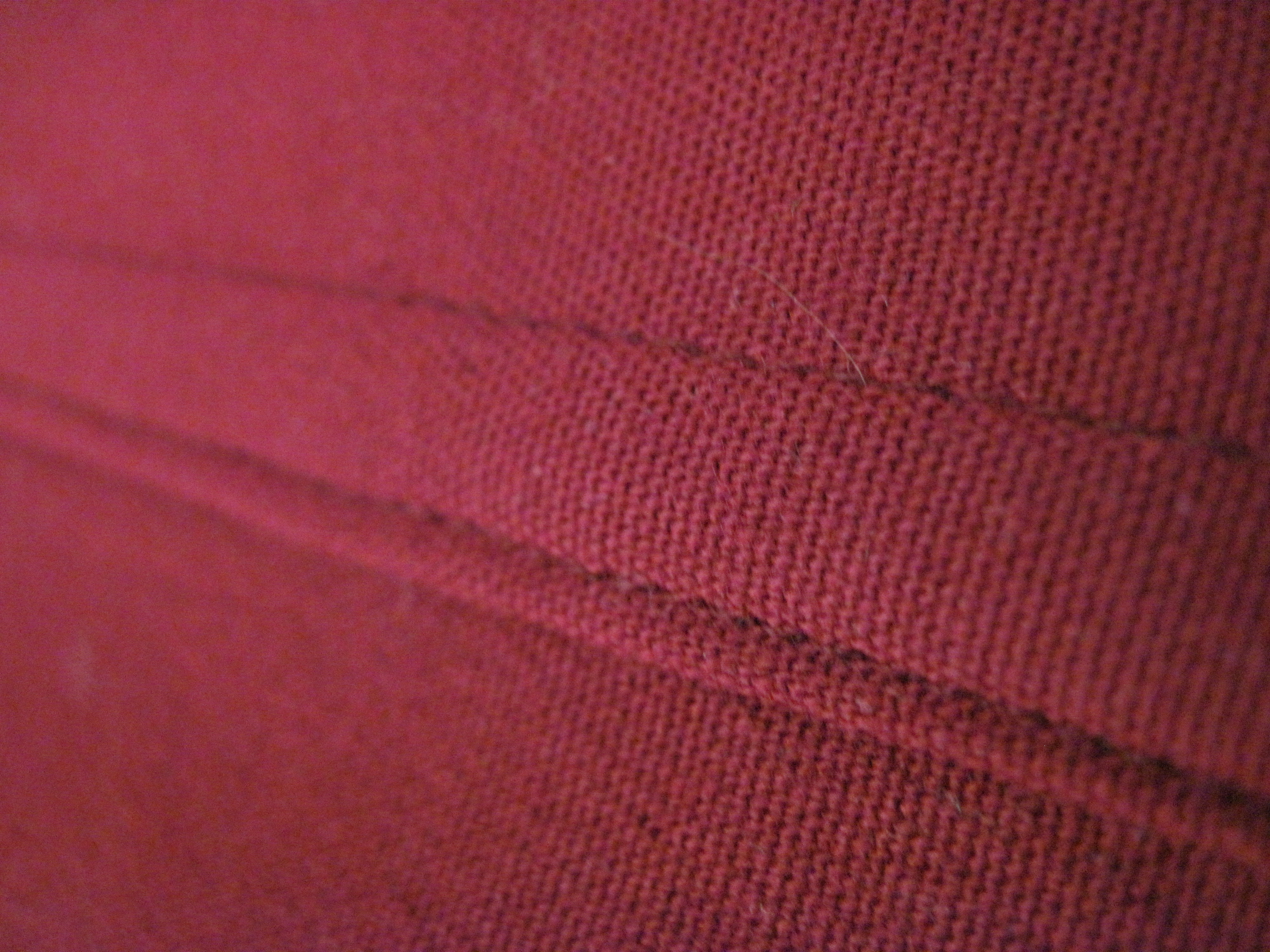 Real photograph of a seam surface and visible stitches on red fabric, identified by its source as flat-felled. Local appearance only; hidden cut edges, stitch length, composition and strength are unverified.